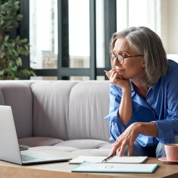 Mature lady looking at the laptop screen with pen in hand and notebooks and coffee on the table