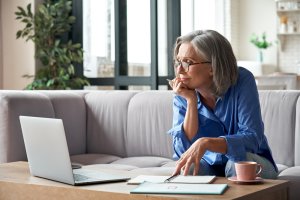 Mature lady looking at the laptop screen with pen in hand and notebooks and coffee on the table