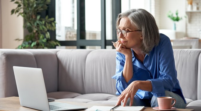 Mature lady looking at the laptop screen with pen in hand and notebooks and coffee on the table
