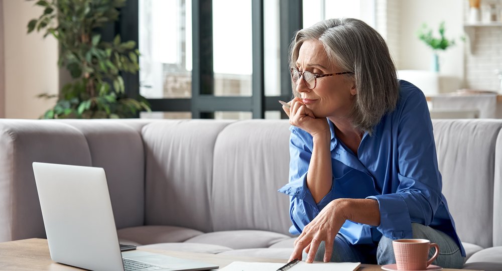 Mature lady looking at the laptop screen with pen in hand and notebooks and coffee on the table